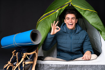 Teenager caucasian man inside a camping green tent isolated on black background surprised and showing ok sign