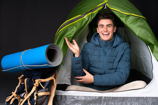 Teenager caucasian man inside a camping green tent isolated on black background extending hands to the side for inviting to come