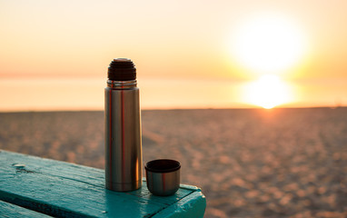 Thermos with fragrant hot tea on a blue bench, on the shore by the sea. Sandy deserted beach.