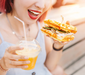 Smiling hipster woman drinks fresh juice and eats a delicious sandwich with a loaf of a checkered Viennese waffle.