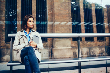 Thoughtful young woman dressed in stylish coat looking away holding modern touch pad device in hands.Pondering hipster girl with tablet sitting on tram stop waiting on transport in urban setting