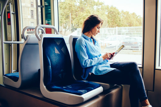 Hipster girl sitting in tram using modern touchpad for chatting in social networks with friends via free wifi connection, young woman reading e-book downloaded on portable pc in public transport.