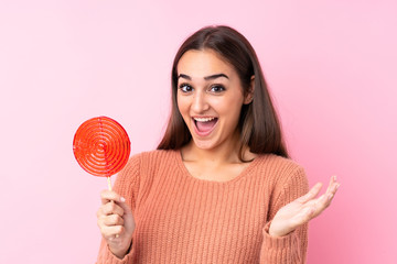 Young girl holding a lollipop over pink background with shocked facial expression