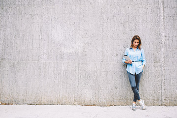 Stylish young woman holding modern touch pad device in hands and looking down.Casual dressed hipster girl with tablet standing near promotional background with copy space area for your advertisement