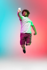 Fototapeta na wymiar Phone scrolling. Caucasian young man's portrait on gradient studio background in neon. Beautiful male model in casual style jumping high. Concept of human emotions, facial expression, youth, sales, ad