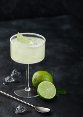 Luxury glass of Margarita cocktail with fresh limes and bar spoon with ice cubes on black background.