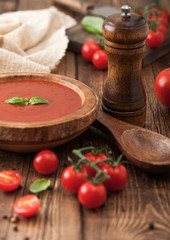 Wooden plate of creamy tomato soup with wooden spoon, pepper and kitchen cloth on wooden background with raw tomatoes.