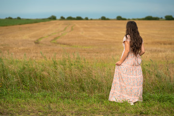 Fototapeta na wymiar Beautiful young girl walking in a field. A girl in a rustic dress with long hair stands with her back to the camera and looks at a freshly cut and harvested field. 