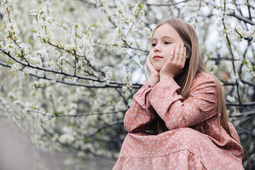 A little girl child sits in the garden under a flowering tree, resting her head on her hands and looking into the distance with a thoughtful look.