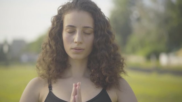 Close-up portrait of confident brunette curly-haired woman putting hands together, closing eyes, breathing in, and looking at camera. Portrait of Caucasian yogi training outdoors in summer morning.