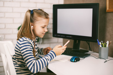 A girl in headphones with a phone in her hands sits at home at the table at the computer and does her homework. Online education. The child learns from home. Distance learning online.
