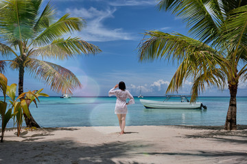 A girl looking horizon standing next to a palm trees on a beach in front of a Caribbean sea