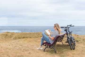 Fototapeta premium Woman sitting on a bench in front of the sea reading a book and two bikes leaning against the bench.