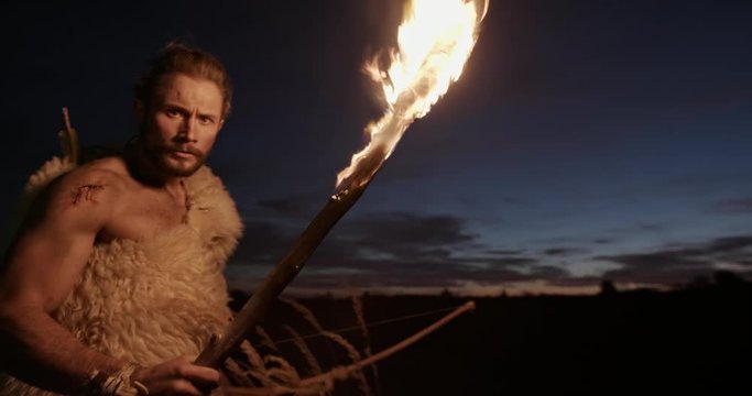 Male hunter in animal hide carrying burning torch and looking at camera suspiciously while exploring wilderness at night. Wild aborigine, savage or Neanderthal Viking on a sunset background.