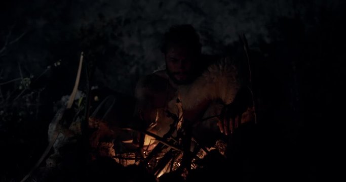 Bearded man in animal hide adjusting firewood and putting apple in campfire while resting and cooking at dark night in wilderness. Reproduction of the historical life of Neanderthal man, or Cro-Magnon