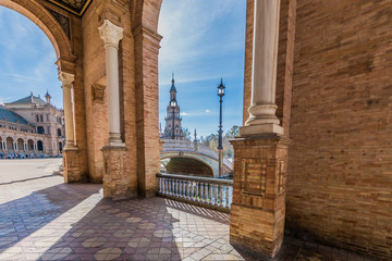 Corridor with pillars and arched openings in Plaza de Espana with part of the building, pole lamps and the bridge in the background, sunny day with a blue sky in Seville, Spain