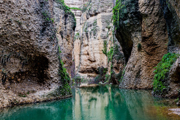 Huge rocky mountains with moss and plants between its slopes, the Guadalevin river with its calm turquoise waters with reflection, sunny day in Ronda, Malaga province, Spain
