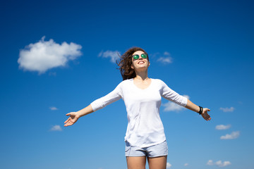 Free happy young woman in white clothes and sunglasses with arms outspread and face raised in sky, hair fluttering in the wind, enjoying life and sunny day, standing on blue cloudy sky background