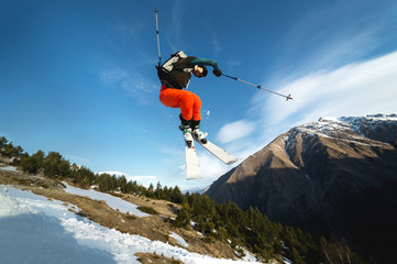 A young stylish man in sunglasses and a cap performs a trick in jumping with a kicker of snow against the blue sky and the sun on a sunny day.
