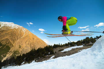 Girl skier in flight after jumping from a kicker in the spring against the backdrop of mountains and blue sky. Close-up wide angle. The concept of closing the ski season and skiing in spring - obrazy, fototapety, plakaty