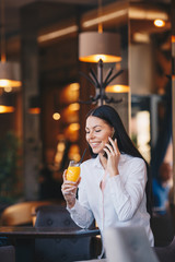 Young woman is sitting at a table in a pub, talking on her mobile phone and drinking orange juice
