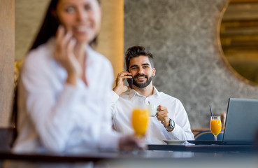 A young woman is sitting at a table in a restaurant, talking on a mobile phone and drinking orange juice, behind her sits a young gentleman and talking on the phone