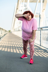 An obese woman does a warm-up exercise on the bridge and wipes the sweat from her forehead with her hand