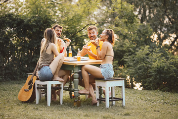 Two couples sit outside in nature, enjoying themselves, eating pizza and drinking beer