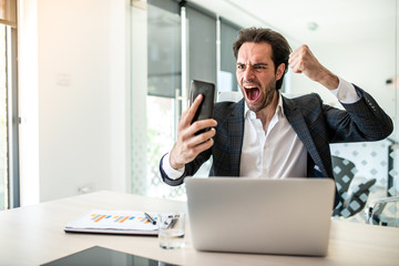 Image of a young businessman sits at a desk in the office, holds a mobile phone in his right hand...