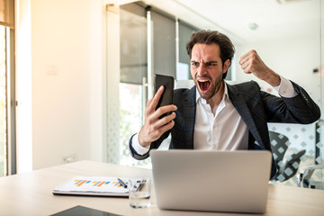 Young businessman sits at a desk in the office, holds a mobile phone in his right hand and rejoices