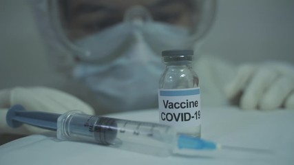 human wearing protective PPE kit approaching and slowly grabbing covid 19 vaccine placed near syringe.