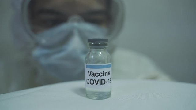 doctor wearing protective PPE kit slowly approaching toward an isolated covid 19 vaccine from a blurred background.