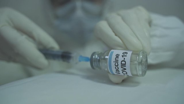human wearing protective PPE kit carefully initiating to draw out covid 19 vaccine from vial into syringe in horizontal position.