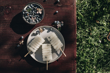 Vegetarian ice cream. On the plate are three white fruit ice creams on a stick. Next is a bowl with blueberries and flowers. Wooden background green grass is visible from the side. Fruit ice cream.