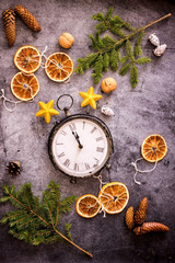Christmas clock composition. Vintage clock surrounded by fir branches, cones, dried oranges, Christmas tree decorations on a dark background. Top view, copy space, flat lay