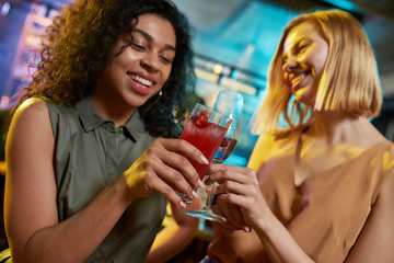 Party On. Attractive young women looking cheerful while toasting, posing with cocktail in their hands. Friends celebrating, having fun in the bar