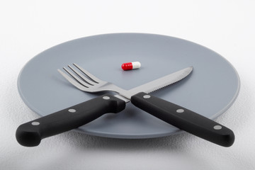 Red-white medical capsule on a gray plate next to a knife and fork on a white background. Medication intake concept.