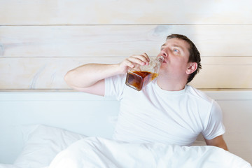 Drunk rowdy man with angry face, bottle of alcohol sits drinking in bed in wooden bedroom. Man with...