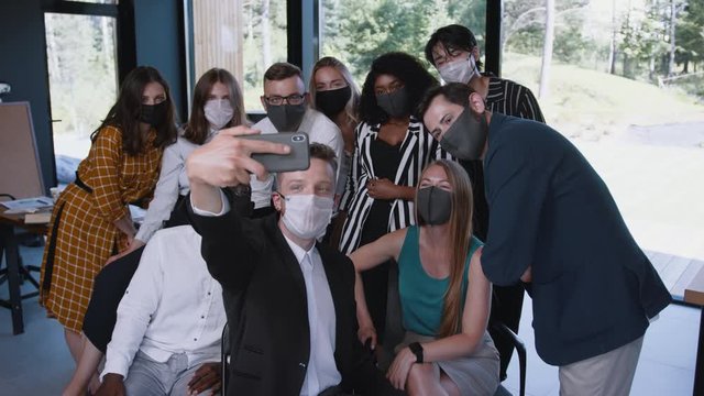Business after COVID-19. Team of happy young diverse colleagues waving for selfie photo in safety face masks at office.