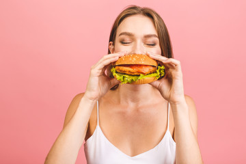 Portrait of young beautiful hungry woman eating burger. Isolated portrait of student with fast food over pink background. Diet concept.