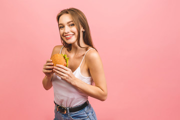 Portrait of young beautiful hungry woman eating burger. Isolated portrait of student with fast food over pink background. Diet concept.