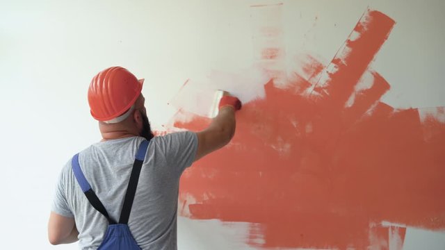 A bearded male construction worker is dressed in overalls and a protective helmet. paints the wall with white paint. Removes stain. Renovation concept in the room