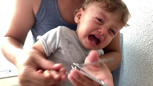 Tearful baby crying while mother trims hand nails. Parent trimming toddler nail