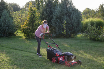 Tired woman making a short break while cutting grass with a lawn mower. Outdoor household chores concept.