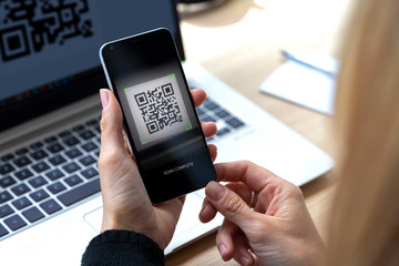 Woman with a cell phone in her hand scanning a QR code from the screen of laptop.