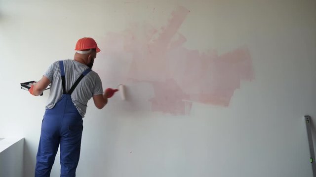 A bearded male construction worker is dressed in overalls and a protective helmet. paints the wall with a roller, general plan. Renovation concept in the room