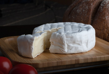 Fresh French Brie cheese and a one slice on a bamboo board with cherry tomatoes, bread on the black plate.