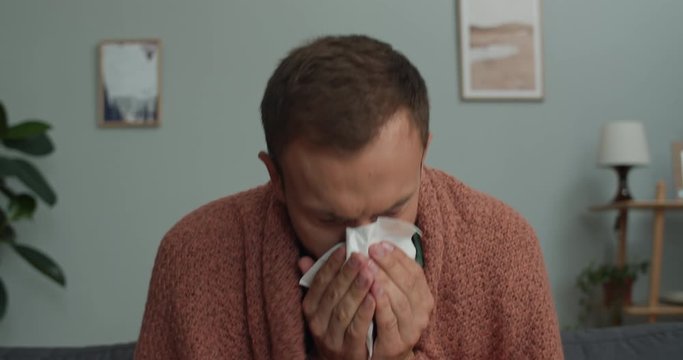 Close up view of young man taking handkierchief and sneezing while sitting on sofa at cosy room. Bearded guy getting sick and spending time at home. Concept of illness and flu
