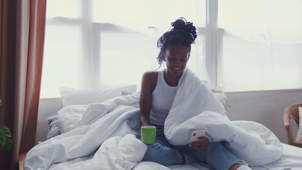 Attractive Young African American Woman sits in bed, drinks morning coffee and uses phone