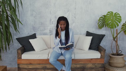 Young african american woman sits sofa, talks on phone with book in hands